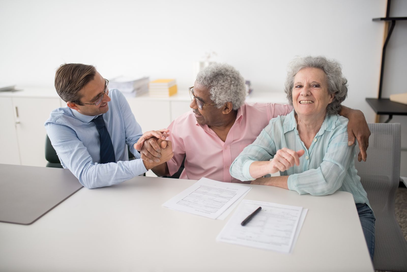 An older couple at a white table with a man in a shirt and tie, papers and a pen in front of them