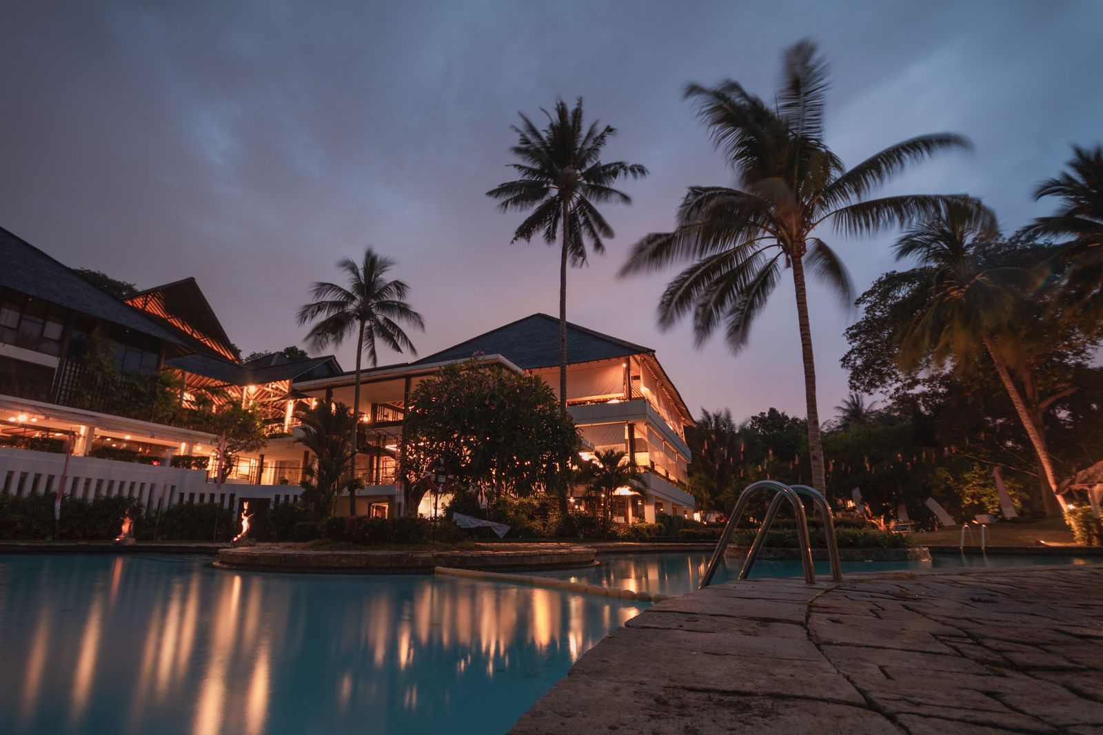 A lit resort building and palm trees reflected in a pool at dusk